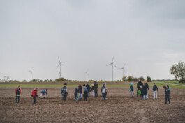 Mehrere Kinder stehen auf einem Acker und bearbeiten ein Bühfeld, im Hintergrund sind Windräder zu sehen